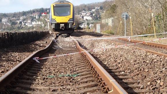 Looking up from train tracks to a carriage which has derailed on its way to Barrow-in-Furness, in Cumbria. The train is Northern operated, with blue and yellow branding. The train tracks have collapsed into a hole in the ground.