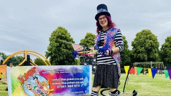 Performer with long purple hair wearing a black top hat and glasses, a dark skirt and black and white top with a multicoloured waistcoat. The performer is holding some coloured plastic straws and is standing next to a banner advertising "the Joker". There is a unicycle behind the performer, some coloured bunting and trees in the background.
