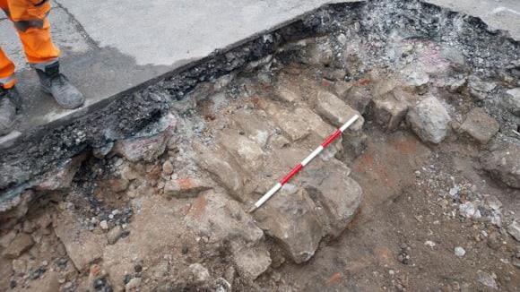An image of a large hole dug into a concrete road, exposing rubble and bricks below the surface. A red and white striped stick marks out a section of the exposed bricks. To the left, stood on the concrete a pair of legs can be seen, wearing black work boots and florescent orange trousers.