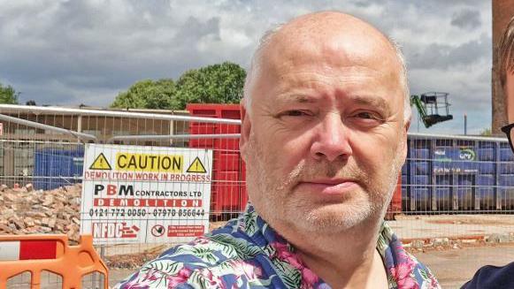 A man with short grey hair and a floral blue shirt standing in front of a building site
