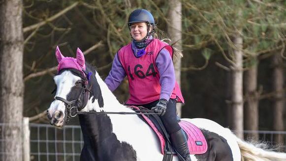 Lucy White is wearing a lilac jumper, pink vest and a helmet. She is riding a horse with a brown and white coat in front of a row of trees.