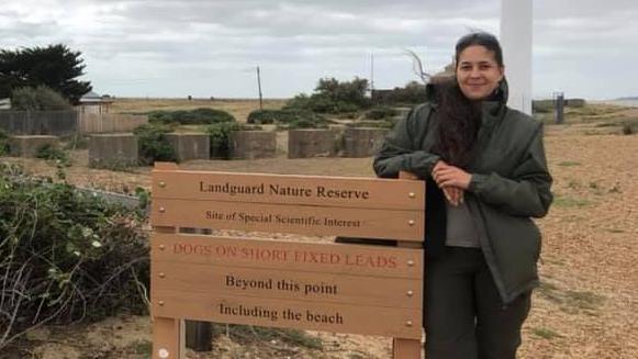 Ms Washington leans against a wooden information board at Landguard Nature Reserve. Her hair is partly tied back and partly down and she wears a green coat with a scarf and black trousers.