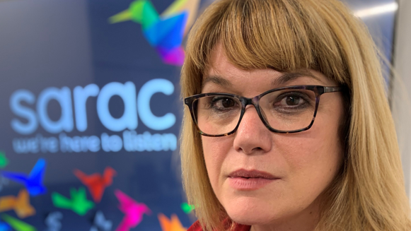 A woman with shoulder length blonde hair and dark framed glasses looks at the camera