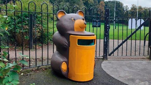 A yellow bin, which is shaped as a bear holding a barrel. There is a fence with a gate in the background. The bin is sat on a tarmac path.