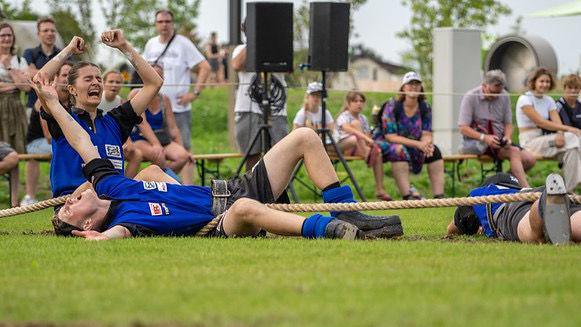 Two tug of war competitors lie on the floor beside their rope. One is faced down, the other is faced up with his hands in the air in celebration. Another competitor kneels with her arms in the air, appearing to shout with joy.