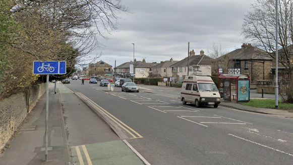 Google streetview image of a B-road.  Vans and cars are travelling towards the direction of the camera. There is a cycle lane on the left and a park on the left side of the pavement.
