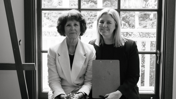 A black and white photo of Cherie Reid and Karina Reid sitting next to each other on a window sill. They are both looking at the camera and smiling.