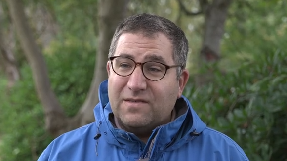 Rob Kantor during a TV interview. He wears dark glasses and a blue coat as he stands in front of bushes and trees.