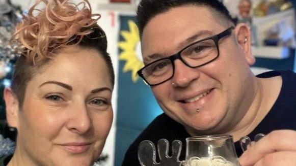 Clair Honeywood with her partner Danny Burch. She has short brown hair with a spiral of pink curls on top. Her partner has dark hair and piercings in his nose, lip and ear.
