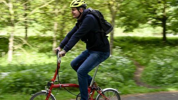 A man wearing a yellow bike helmet rides a red city bike. The background is trees and shrubbery but is blurred because of the travelling cyclist.