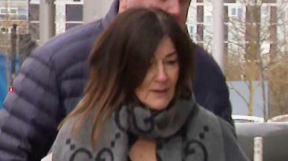 A woman with dark hair and a grey and black scarf looks down. She appears to be outside and a man wearing a navy puffa jacket is behind her.