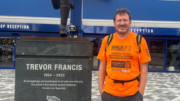 Xavier Wiggins is dressed on the right in bright orange top that says Walk 92. He's stood next to the Trevor Francis memorial bronze statue outside St.Andrews at Knighthead Park.