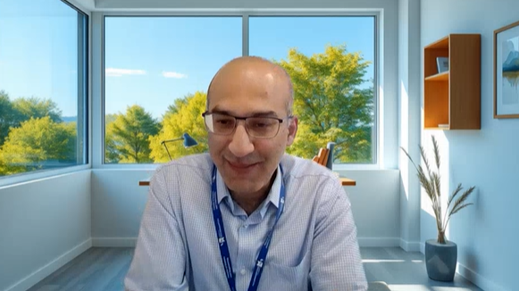 Mr Pervez is sat down with a fake office background image behind him during an interview. He is wearing glasses, a blue shirt and has closely cropped grey hair.