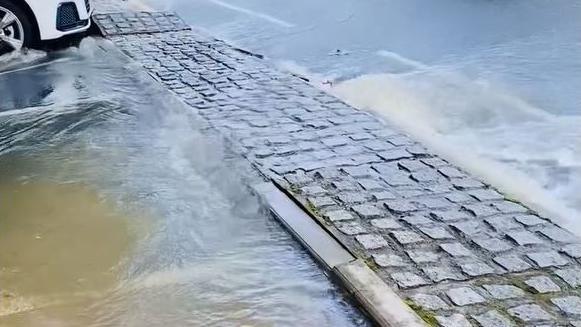 A large flow of water overflowing from drains beneath a cobbled path adjacent to a road.