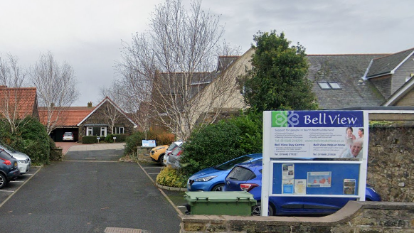 A sign saying Bell View is in the foreground of the picture and behind you can see a modern one storey building. There is a driveway in front with parking spaces in either side  