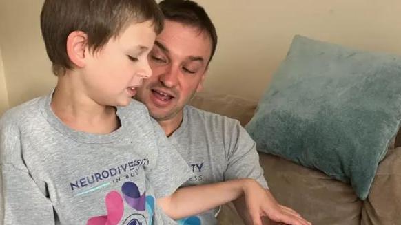 Joshie sitting on his dad's lap, both are wearing grey T-shirts and black trousers