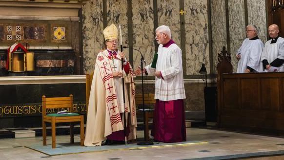 John Sherrington wears an ornately decorated cream and gold mitre and a white and red robe in a cathedral. A men in a maroon and white robe stands beside him.