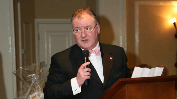 A man with grey hair and glasses stands at a lectern, holding a microphone, and turning to look towards the left. He wears a light pink bow tie, white shirt, and black suit jacket. He is in a room with cream-coloured walls and white doors.