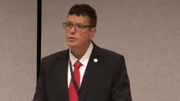 MS Nigel Williams in the Senedd. He has short dark hair and glasses and wears a  black suit jacket, white shirt with a red tie. 