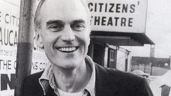 A black and white shot of a man outsizxe the Citizens' Theatre in Glasgow. He is clean shaven with short hair and has a wide smile. He wears a dark blazer, a shirt unbuttoned at the neck and a light t-shirt, with a Citizens' Theatre badge on his lapel.