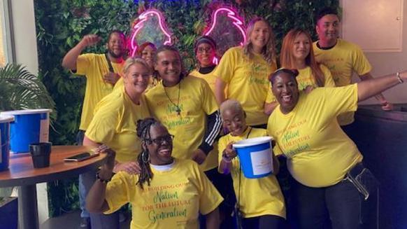 Cheering bar staff wearing yellow T-shirts hold a blue bucket for donations, in front of a green, leaf-motif wall at Mavericks Bar