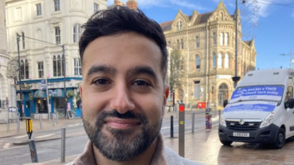 A man with short black hair and a short dark beard smiles at the camera. He is standing outside on a street, two sandstone buildings can be seen behind him, and on the right is a white van.