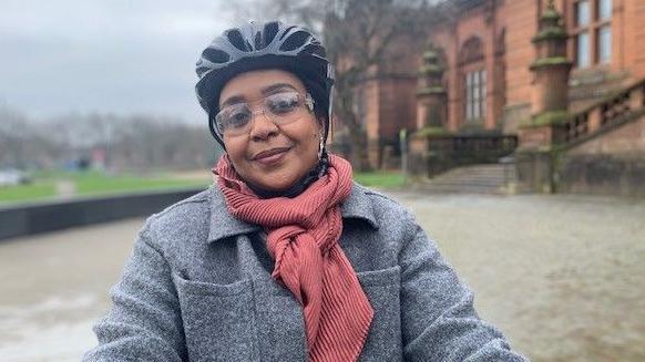 Nahla Abdulla sits on her bike outside the imposing red sandstone building that is Kelvingrove Art Gallery. She looks happily at the camera, wearing her black cycle helmet, grey wool coat and a peachy pink scarf.