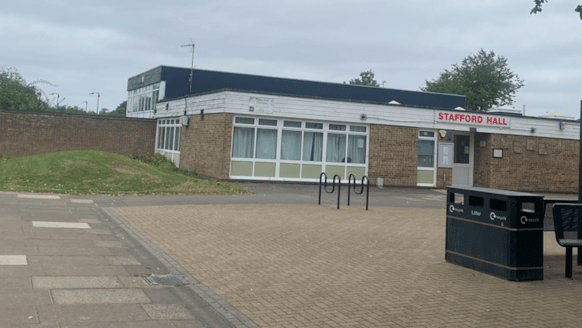 A redbrick modern building has large windows and white wooden cladding across the top. Above the door is a red sign saying Stafford Hall in upper case letters. There are bicycle racks outside and a bin and benches on a pave area in front.