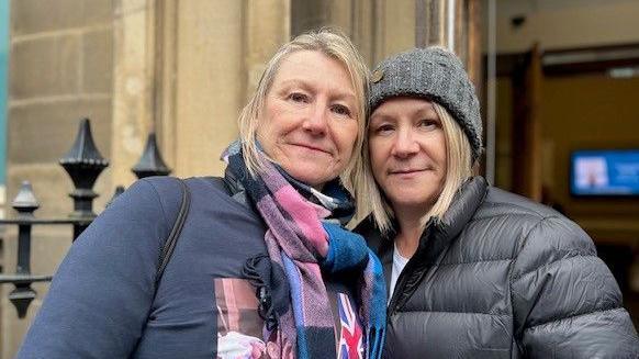 Two ladies with dark padded jackets pictured outside the Town Hall in Huddersfield. The lady on the right is wearing a checked scarf tied at her neck and the lady on the  left is wearing a grey hat. 