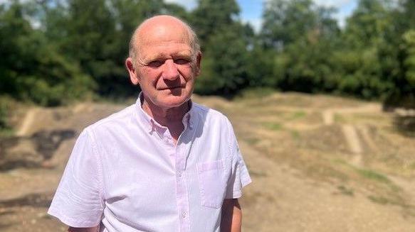A man with a shaven head wears a light-pink short-sleeved shirt. He is standing in front of a dirt bike track with mounds and trails. Green trees stand in the background.