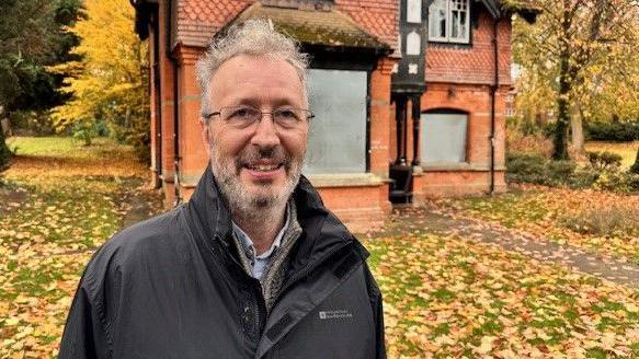 A man in a black anorak is standing in front of a boarded up building in a park. There are autumn leaves all over the ground. He has a beard and he is wearing glasses.