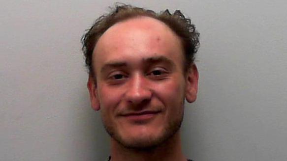 A custody picture of a young man with short, thinning curly hair, who sits against a plain light grey wall, wearing a grey crewneck sweatshirt. He looks directly at the camera with a relaxed posture and a subtle smile.
