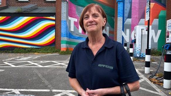A woman in a blue T-shirt and red hair standing in front of a brick building covered in brightly coloured swirls and lines of paint.