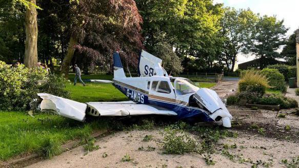 A wrecked white and blue Piper PA-28-140 light aircraft in the middle of a gravel patio large front garden. A man can be seen walking away from the wreckage in the far distance towards large trees.