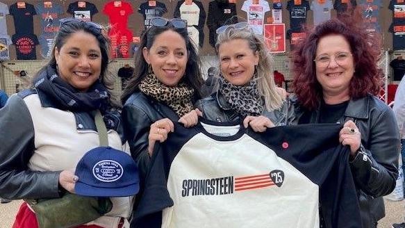 Four women dressed in jackets and scarves stand in front of a Bruce Springsteen merchandise stall holding a Springsteen 25 t-shirt and cap