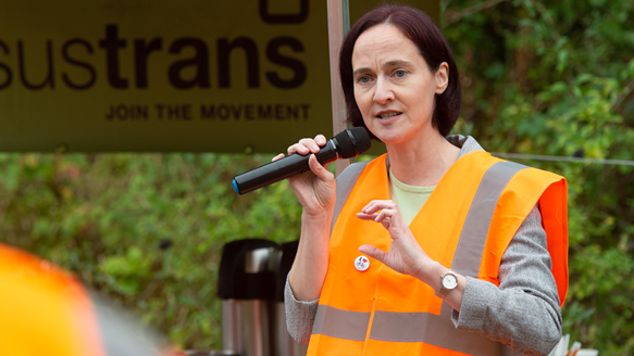 A woman holding a microphone and wearing hi-viz in front of a Sustrans sign against a backdrop of woodland.