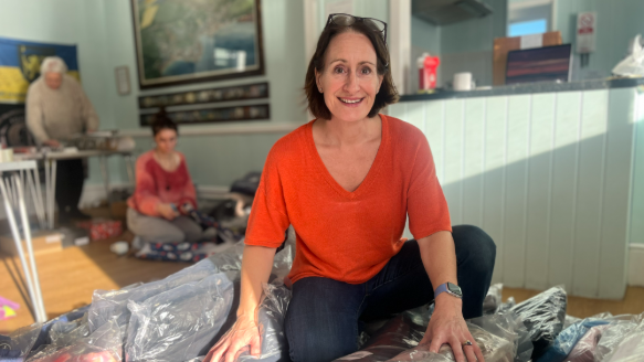 The photo shows Lesley Pond sitting on the floor amongst new clothing in plastic bags. She is smiling at the camera and wearing an orange top. 