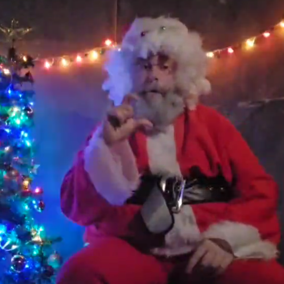 A man dressed as Father Christmas using sign language with Christmas lights behind him and a lit up Christmas tree next to him.