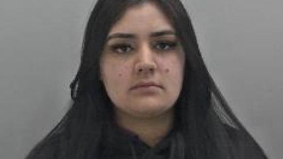 Sanaa Shahzad is a young woman who has long straight, dark hair and is seen wearing a black jumper. She has brown eyes and a mole on each cheek. 