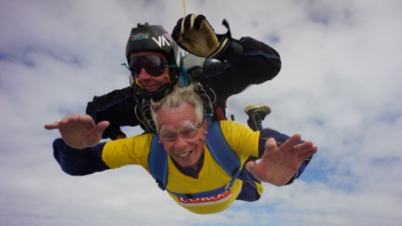 David Hartridge, in a yellow, LOROS t-shirt, smiling widely as he falls from the sky whill harnessed under a skydiving instructor