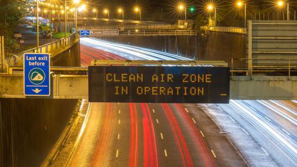 A sign above a motorway saying "CLEAN AIR ZONE IN OPERATION'