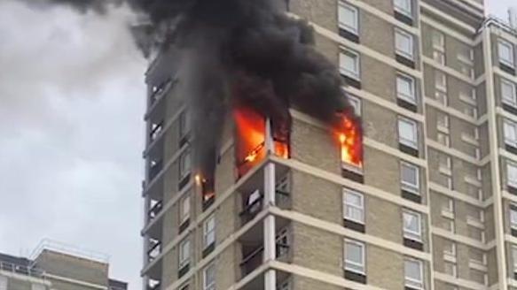 Fire pours out of the windows on two sides of a grey concrete tower block as thick black smoke engulfs the floors above the blaze. One floor of the block is alight. 