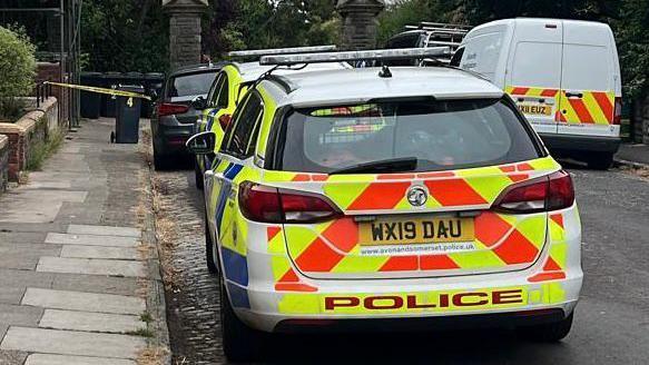 Two police cars and a police van parked on the edge of Victoria Park in Bristol. Police tape can be seen cordoning of an entrance to the park. 