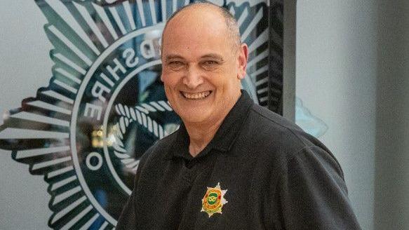 A bald man wearing a black jumper with a fire service badge, smiles while standing in front of a wall with a large fire service badge displayed on it.