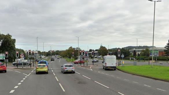A busy junction controlled by traffic lights. A number of cars are waiting at the lights. A retail park is visible to the right of the shot.