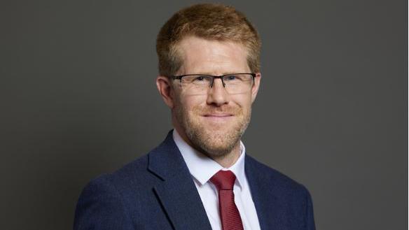 A man with ginger hair wearing glasses, a blue suit, white shirt and red tie smiles at the camera