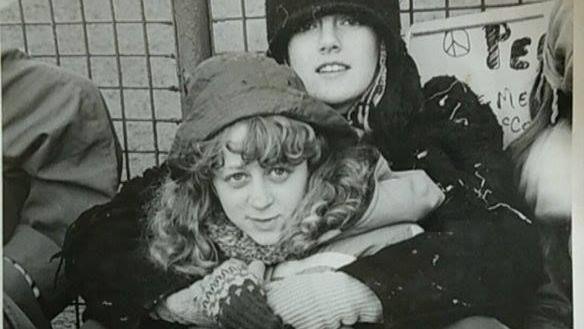 A black and white photo of two women sitting on the ground with their arms around each other. One woman is wearing a black hat. The other woman has curly hair and is also wearing a hat. There are protest posters on a fence behind them, with peace symbols on them.