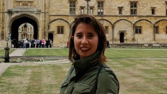 Ms Elena Vento stands by a lawn in front of a historical building. She has shoulder-length, brown hair and wears a green jacket.