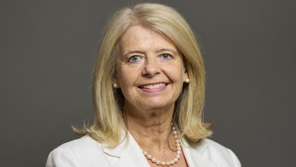 A woman with shoulder length blonde hair, wearing a white jacket and a pearl necklace smiles at the camera