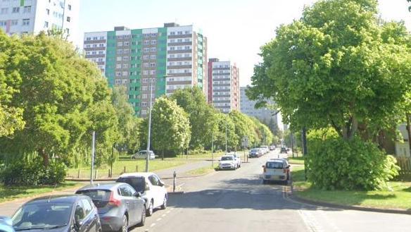 High rise flats along a street with trees and cars parked along it.
